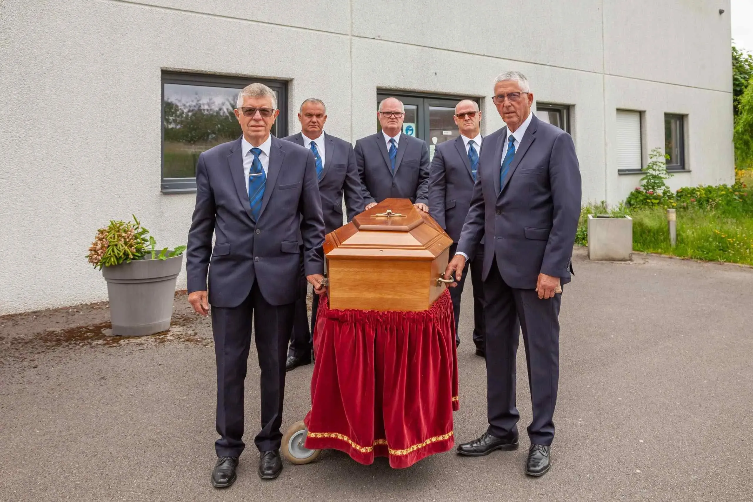 Équipe de porteurs funéraires en costume cravate encadrant un cercueil sur chariot.
