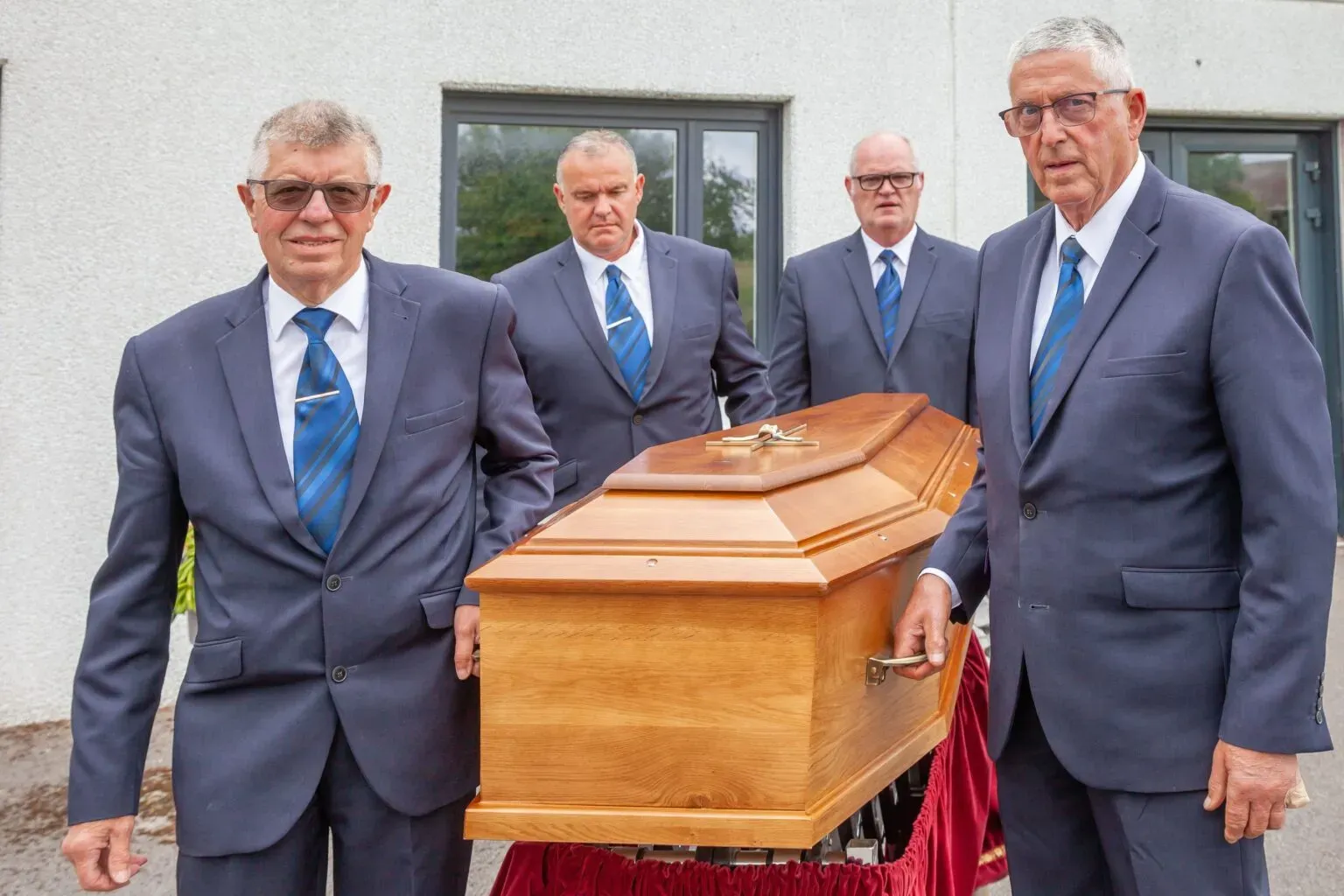Gros plan sur quatre porteurs funéraires en costume guidant un cercueil en bois.